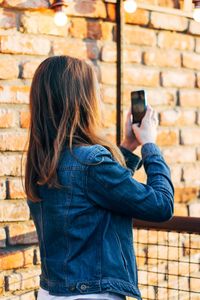 Side view of young woman photographing with mobile phone while standing by brick wall