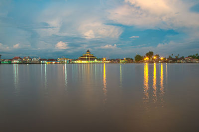 Illuminated buildings by lake against cloudy sky