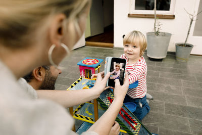 Mother photographing smiling daughter playing with toy cars on porch