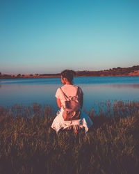 Rear view of woman crouching at grassy lakeshore against clear sky during sunset