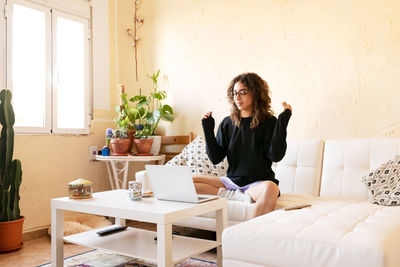 Young thoughtful female freelancer in eyeglasses sitting with eyes closed on sofa and using laptop while working on remote project at home