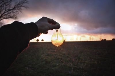 Close-up of hand holding light bulb against sky during sunset