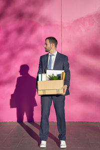 Businessman carrying box with personal belongings in front of pink wall