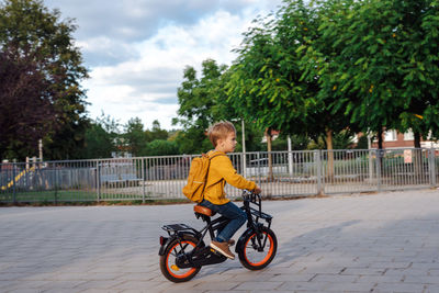 A boy in a yellow sweater with a yellow backpack rides a bicycle in the neighborhood.