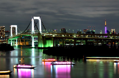 Illuminated bridge over river by buildings against sky at night