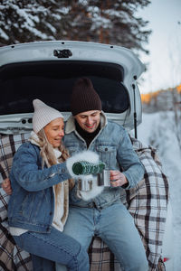 A loving young couple in an embrace sits in the trunk of a white car in winter 