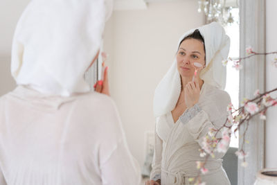 Portrait woman wrapped in white towel and in bathrobe looks at reflection