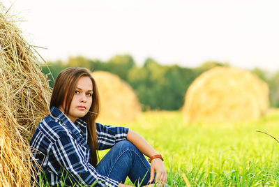 Thoughtful beautiful woman sitting by hay bales on grassy field against sky