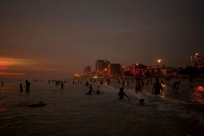 Silhouette people on beach by buildings against sky during sunset