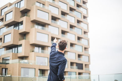 Rear view of man pointing at building while standing by glass railing on terrace