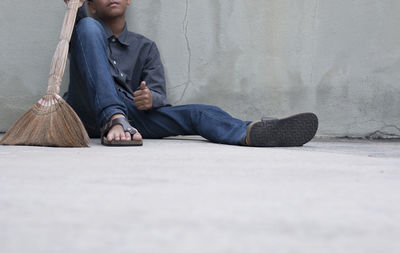 Low section of boy with broom sitting on footpath against wall