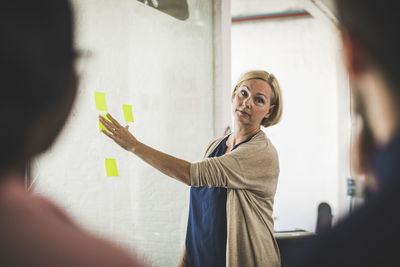 Creative businesswoman explaining colleagues over adhesive note on glass during meeting in office