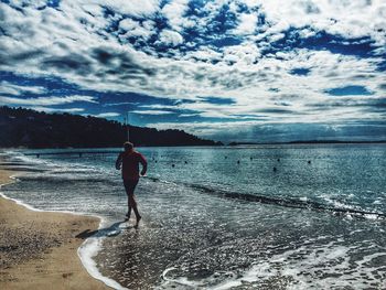 Rear view of man standing on beach against cloudy sky