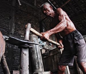 Low angle view of shirtless man working with hammer at workshop