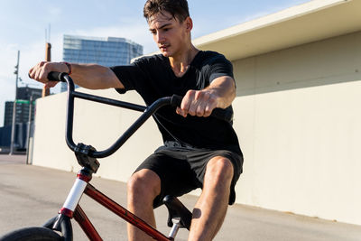 Focused young man in black t shirt and shorts with bmx bicycle near concrete wall on city street