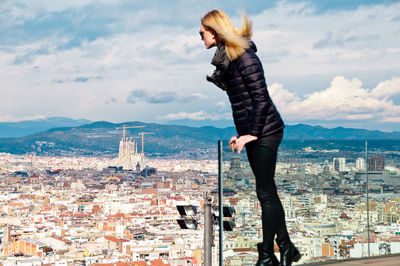 Young woman wearing warm clothing while standing on balcony against cityscape