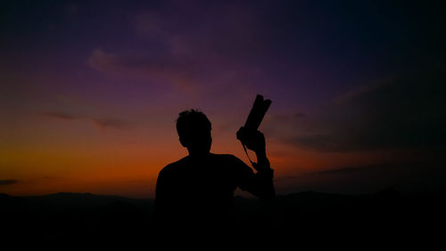 Rear view of silhouette of man holding camera standing against sky during sunset