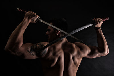 Rear view of shirtless muscular man holding katana swords against black background