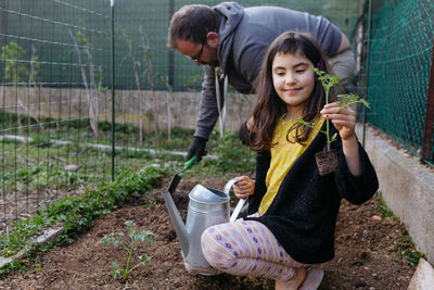 Father digging the garden and daughter with long dark hair holding seedling of tomato in her hand