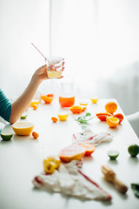 Side view of crop female holding glass of fresh juice with straw while sitting at table with various cut citrus fruits at home