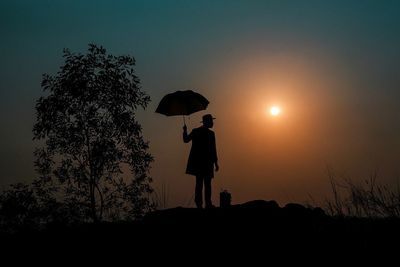 Silhouette man with umbrella standing by tree against sky during sunset
