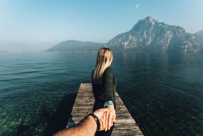 Cropped hand holding girlfriend standing on jetty over lake against sky