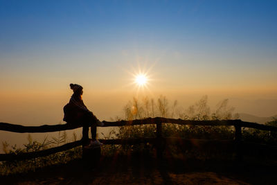 Silhouette woman sitting on fence against bright sun during sunrise