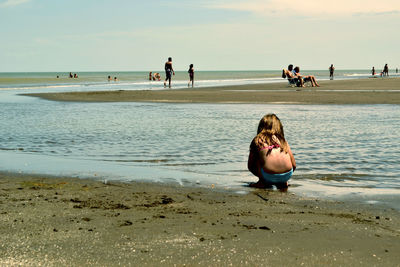 Rear view of girl in swimwear crouching at beach against sky