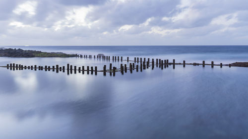 Wooden posts in sea against cloudy sky