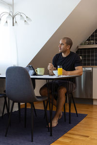 Focused male surfing internet on tablet while sitting at table at home and enjoying breakfast in morning looking away