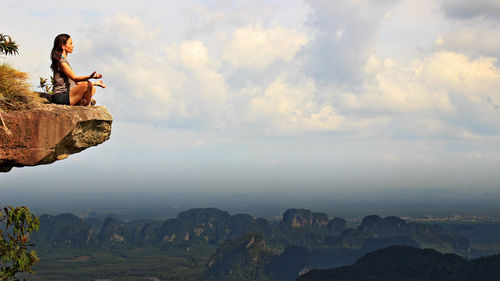 Woman meditating on cliff against cloudy sky