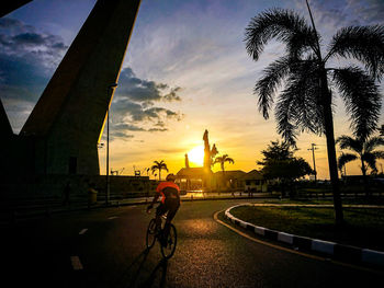 Man riding bicycle on city street against sky during sunset