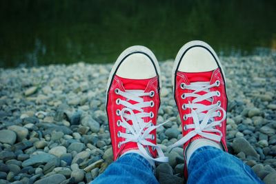 Low section of person wearing red canvas shoe on stones at lakeshore