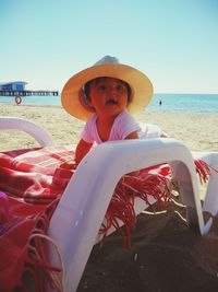 Cute baby girl wearing hat on chair at beach against sky