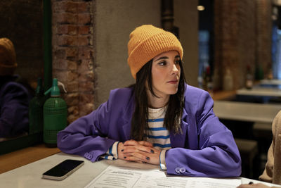 Attractive female in hat and coat sitting at table with menu and smartphone during lunch break in modern light cafe