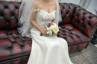 Midsection of bride holding bouquet while sitting on sofa