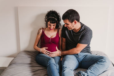 Smiling pregnant woman sitting with male partner on bed at home