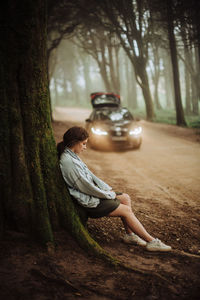 Young woman leaning against mossy tree in forest, car on background