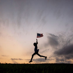 Silhouette girl with american flag running on field against sky