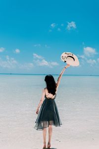 Rear view of young woman holding hat while standing at beach against blue sky during sunny day