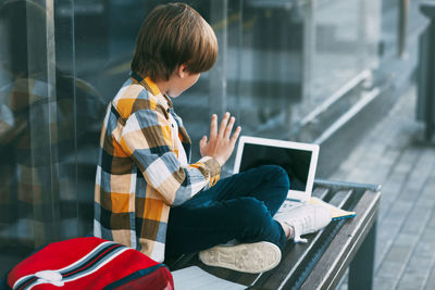 Side view of boy video conferencing over laptop while sitting on bench