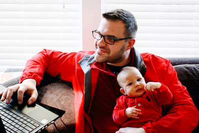 Young man using laptop while sitting on sofa at home holding cute baby in laps 