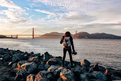 Rear view of woman standing on rocks by sea against golden gate bridge during sunset