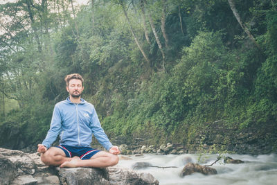 Man doing yoga while sitting on rock at riverbank in forest