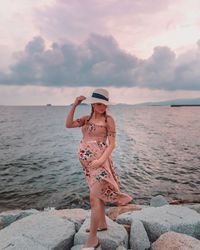 Pregnant young woman touching abdomen while standing at beach against sky