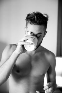 Shirtless young man drinking while using phone at home