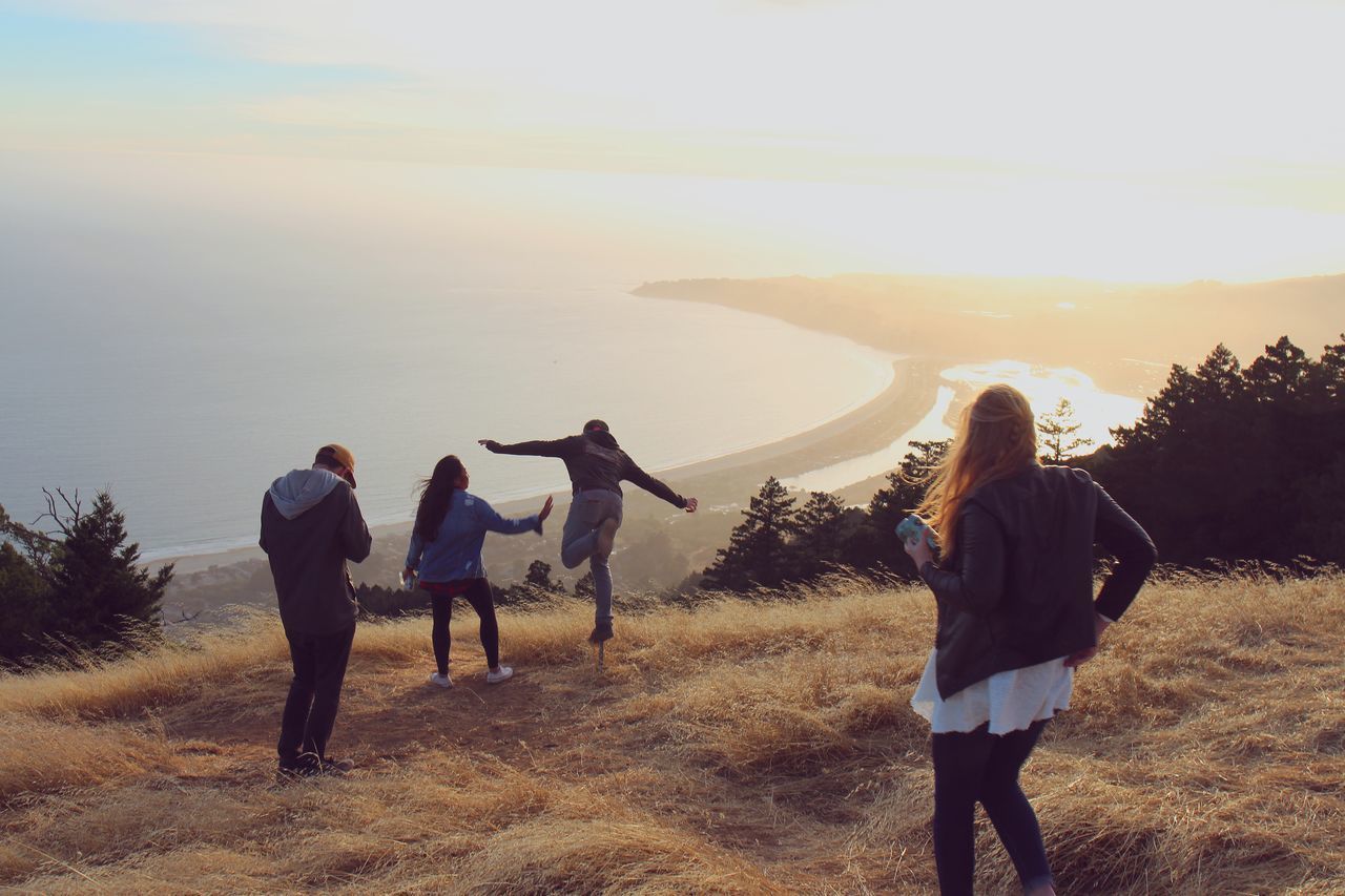 Friends enjoying on grassy hill against sea during sunset