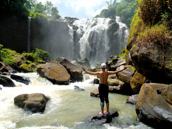 Rear view of man standing with arms outstretched by waterfall