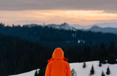 Rear view of man in orange winter jacket looking at snowcapped mountain during sunset