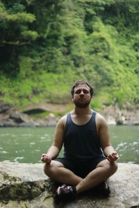 Young man sitting in lotus position on rock against lake